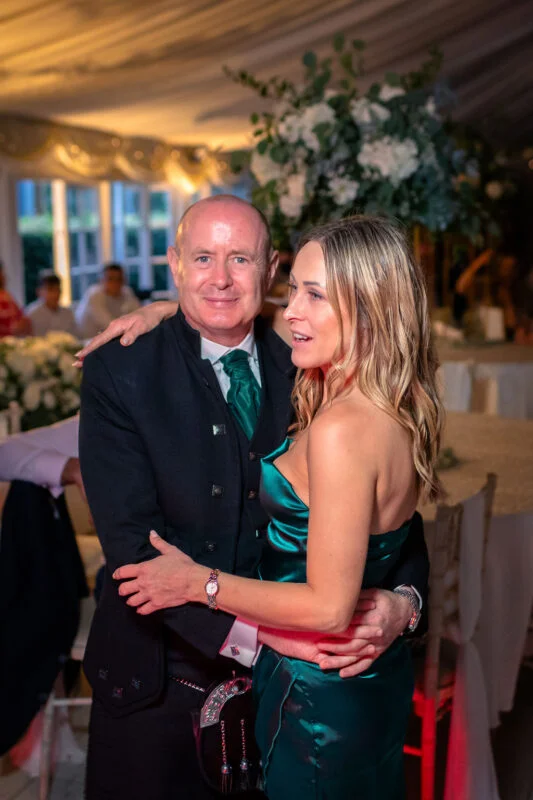 A groom wearing traditional Highland dress with a kilt and green tie embraces his partner in a green satin dress during the evening reception.