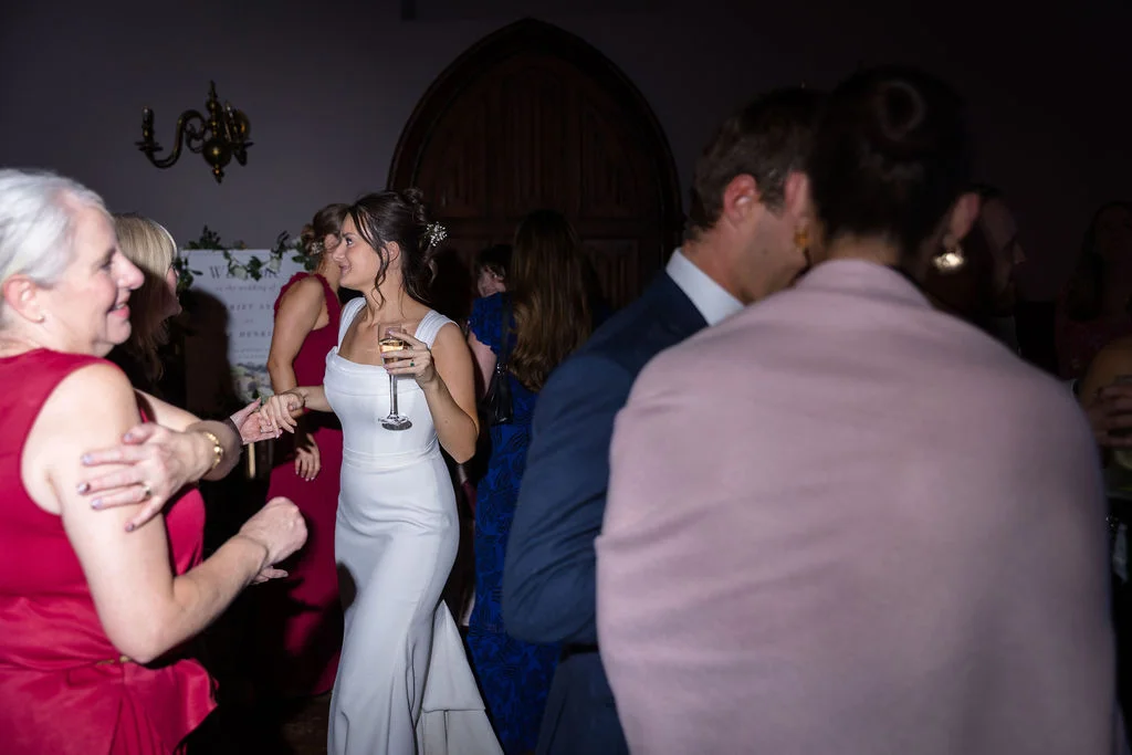 A smiling bride in a white satin column gown holds a champagne glass while mingling with guests at an evening wedding reception.