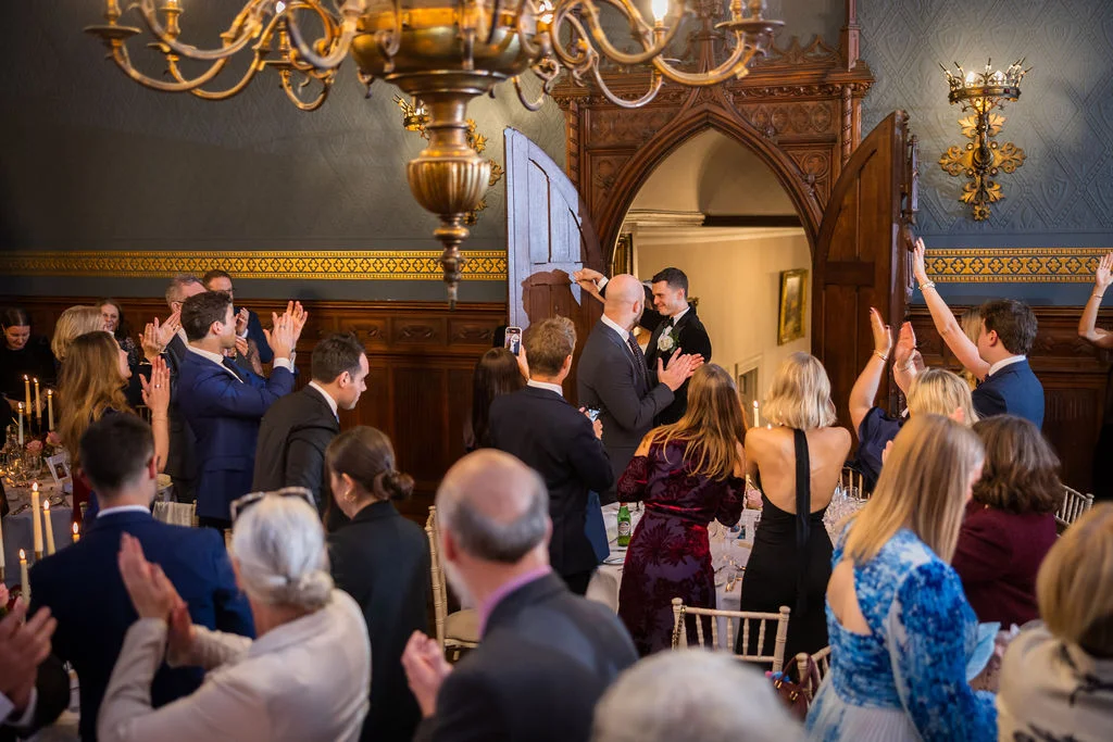Wedding guests stand and cheer as the couple makes their grand entrance into a historic panelled reception room beneath a large ornate chandelier.