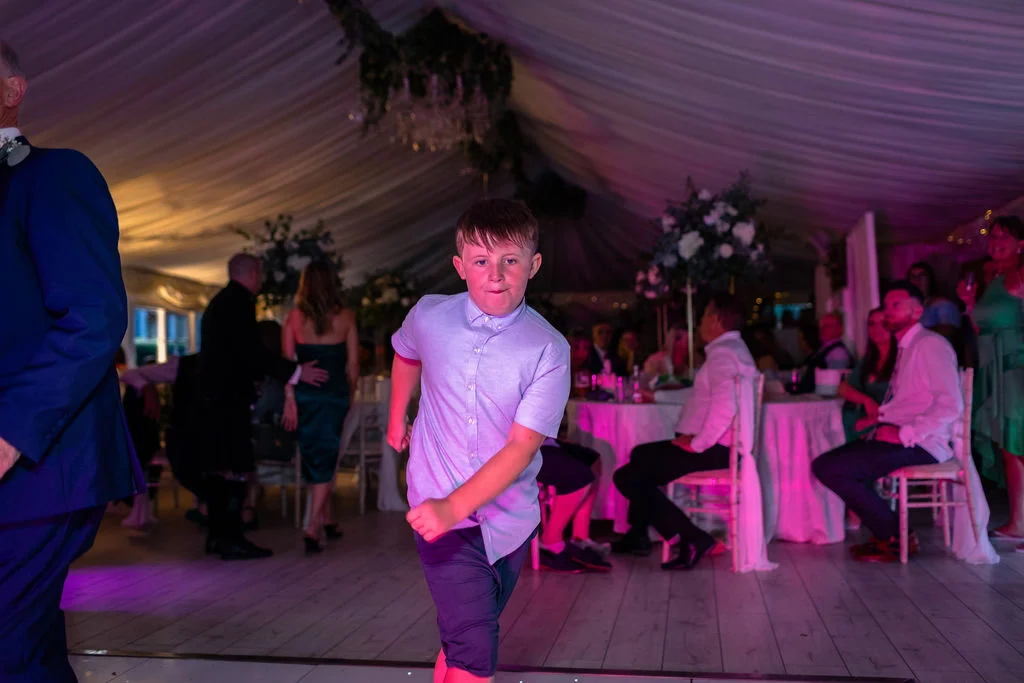 A young boy energetically dances on the lit-up dance floor inside a marquee reception decorated with soft white ceiling draping.