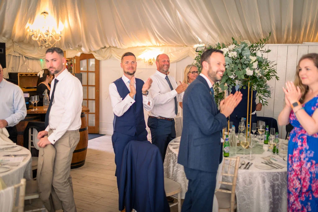 Wedding guests stand and applaud during speeches in a marquee draped with white fabric, featuring round tables with tall greenery centrepieces.