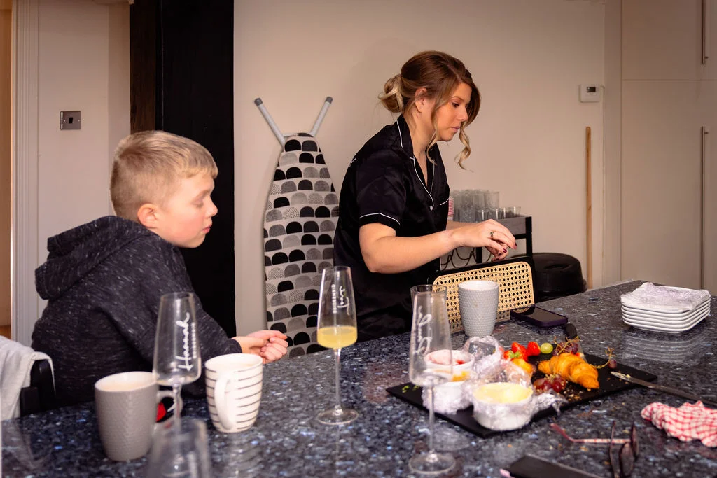 A woman wearing black pyjamas and a low bun prepares a morning spread in a kitchen while a young boy sits nearby.