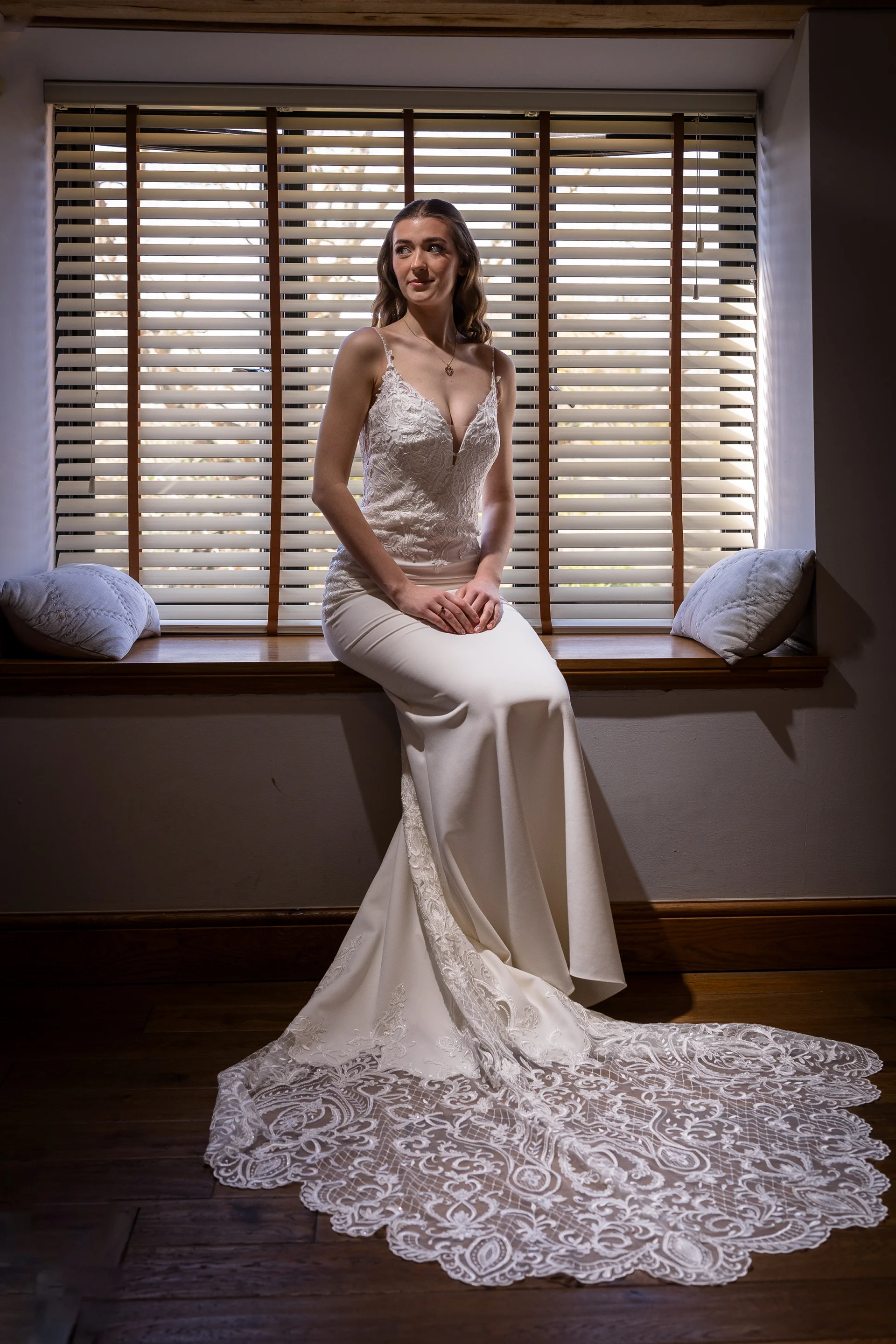A graceful bride sits on a windowsill bathed in soft natural light, showcasing her elegant crepe wedding gown and intricately patterned lace train.