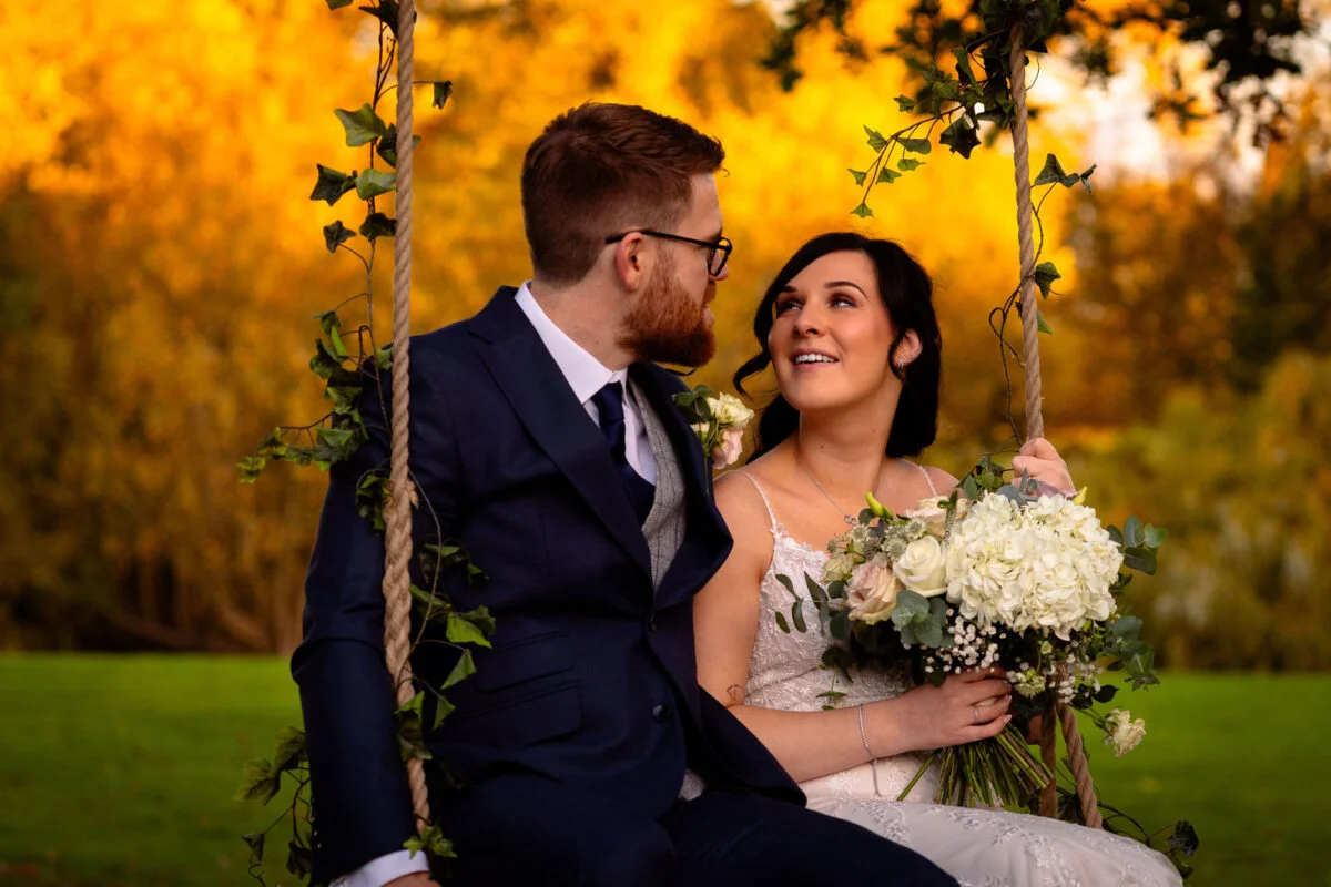 A bride and groom share a tender moment seated on an ivy-wrapped rope swing outdoors amidst vibrant golden autumn foliage.