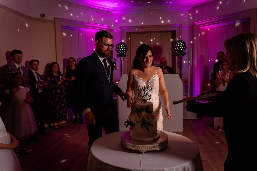 A bride and groom cut their two-tier wedding cake surrounded by guests under vibrant purple disco lighting at a hotel evening reception.