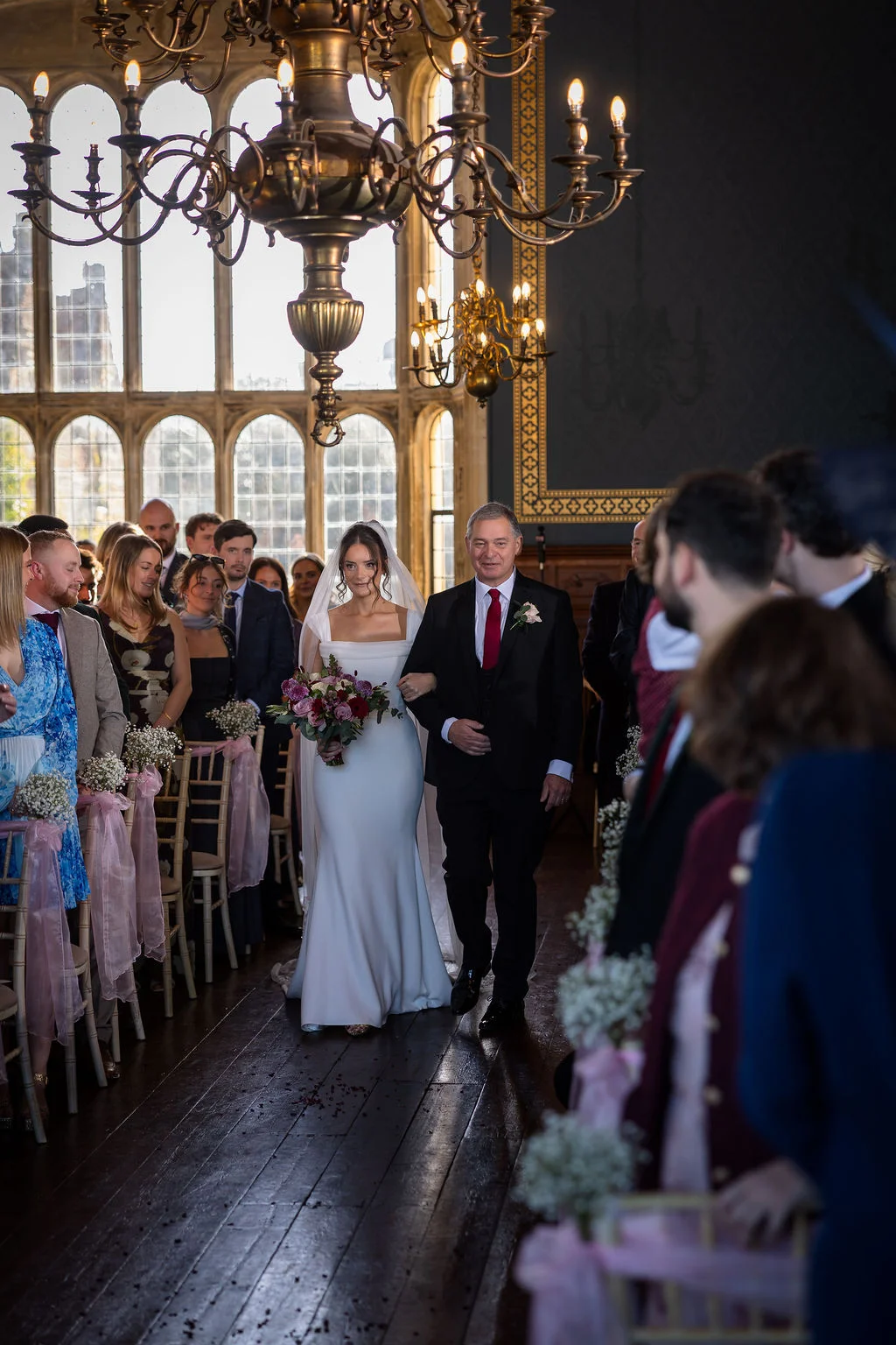 A bride walking down the aisle on her father's arm in a grand hall with high arched windows and chandeliers during a wedding ceremony.