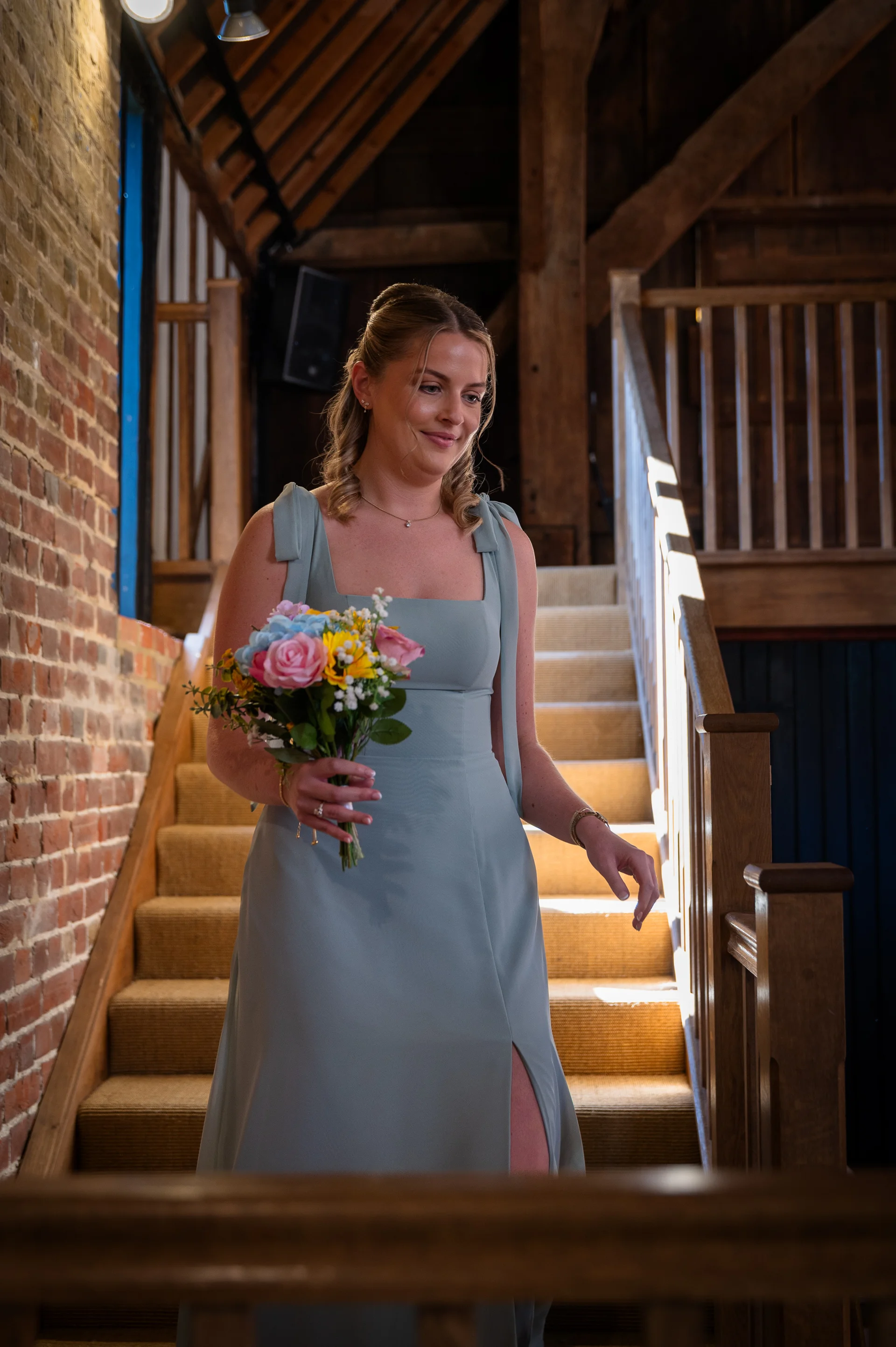 A bridesmaid in a sage green dress holding a colourful hand-tied bouquet walks down a wooden staircase inside a rustic barn venue.