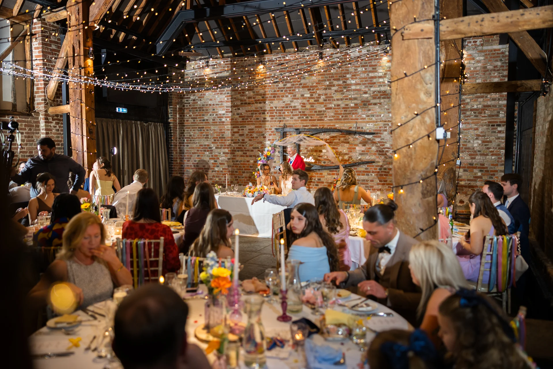 A wedding reception takes place inside a rustic brick barn adorned with a canopy of twinkling fairy lights above the guests seated at round tables.