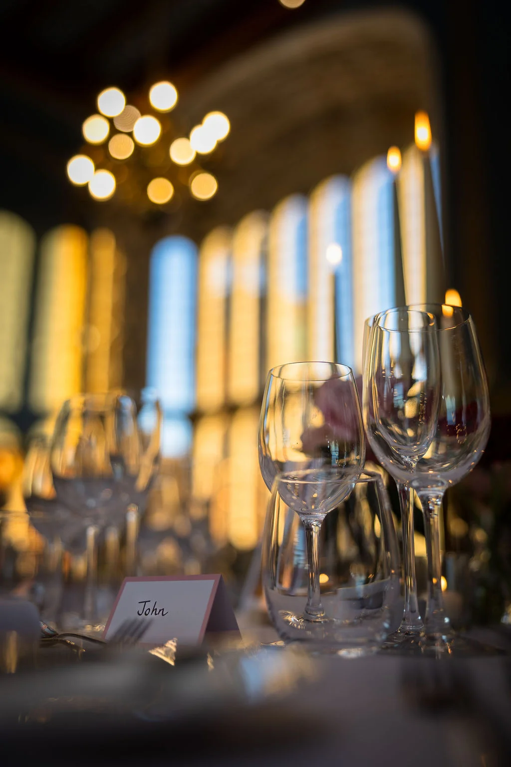 An elegant wedding tablescape featuring wine glasses, a place card bearing the name John, and glowing taper candles against a softly blurred architectural background.