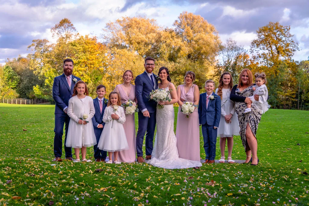 A bridal party poses outdoors on a green lawn scattered with autumn leaves, surrounded by trees with golden foliage.