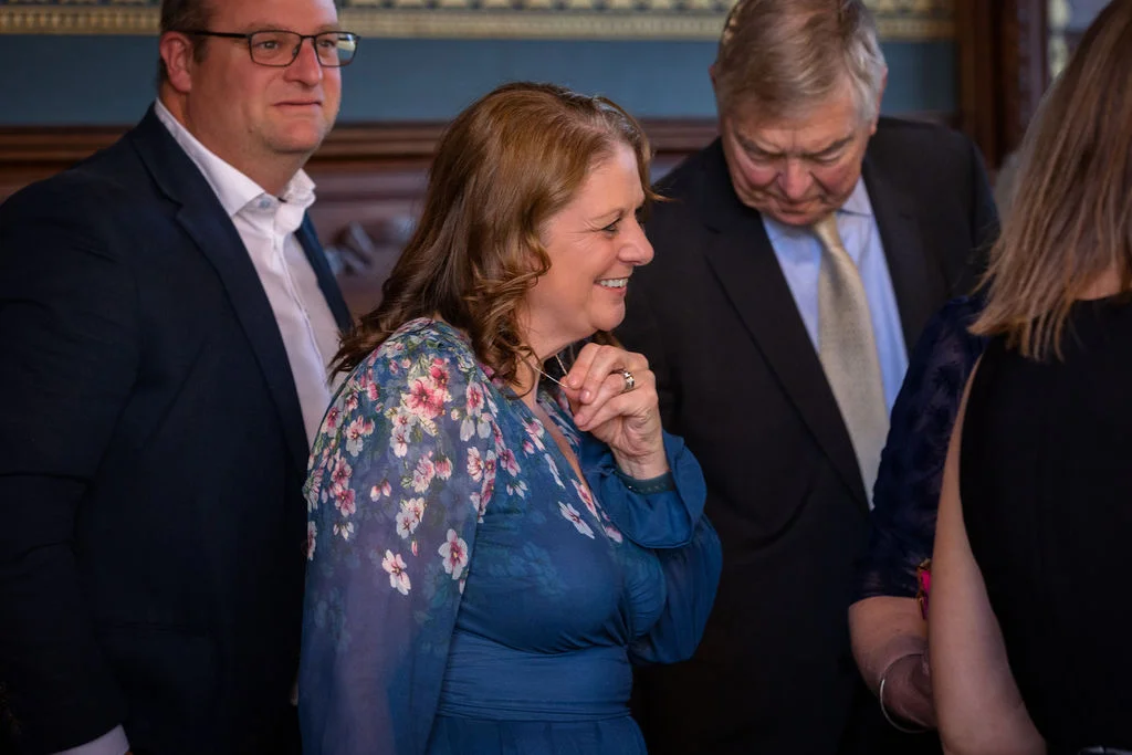 A candid group of wedding guests mingling and smiling indoors, with a woman in the foreground wearing a blue floral dress.