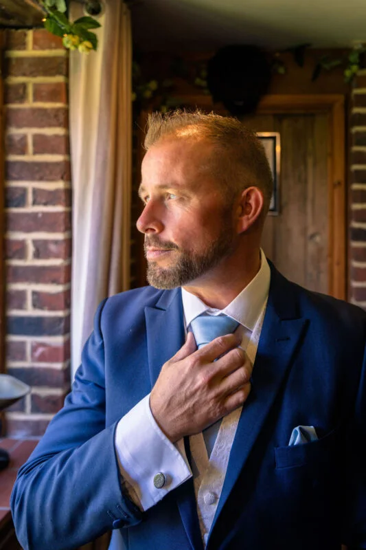 A groom in a blue suit stands by a brick wall, adjusting his light blue tie while getting ready indoors.