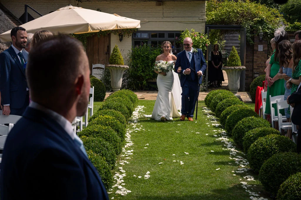 A bride walks down a manicured lawn aisle lined with topiary and white petals, escorted by her father in an outdoor wedding ceremony.