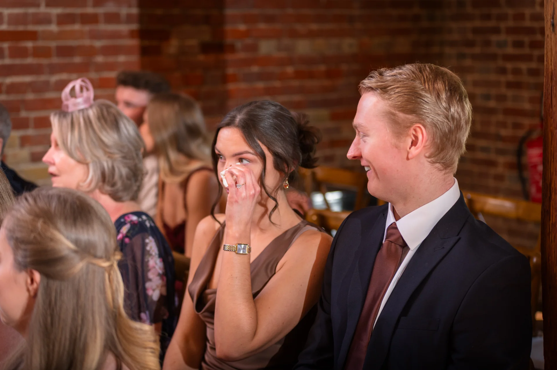 Wedding guests seated in a rustic brick venue are visibly moved during the speeches, with a woman wiping happy tears while a man smiles warmly beside her.