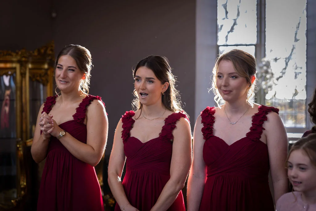Three smiling bridesmaids stand in matching burgundy dresses with ruched bodices and floral straps, illuminated by soft backlight from a window.