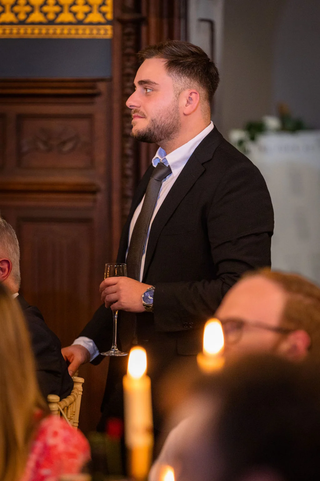 A well-dressed man in a black suit holds a champagne glass while delivering a speech at a wedding reception.