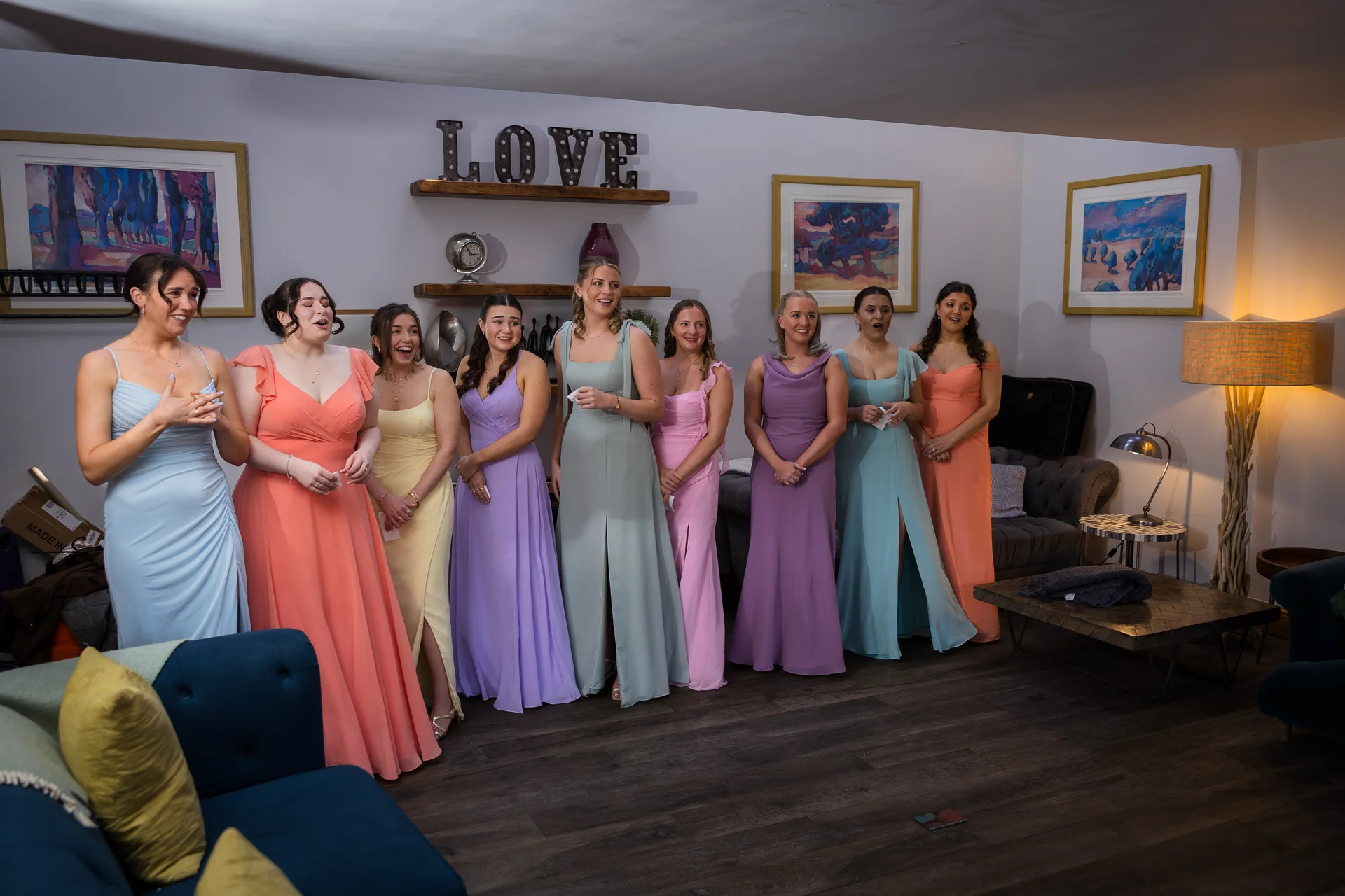 A group of nine bridesmaids in colourful mismatched pastel dresses stand in a row reacting emotionally with laughter and tears during morning preparations.