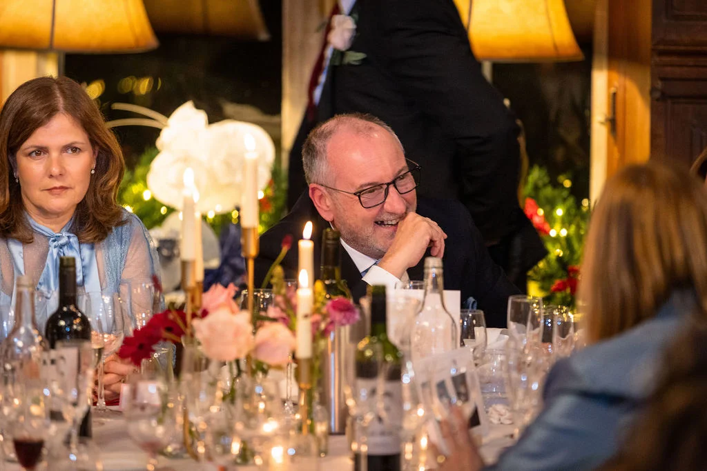A guest laughing heartily during wedding speeches at a candlelit reception table surrounded by wine bottles and floral arrangements.