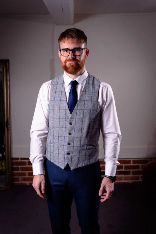 A solo groom with a red beard and glasses stands confidently wearing a grey tweed patterned waistcoat and a blue tie.