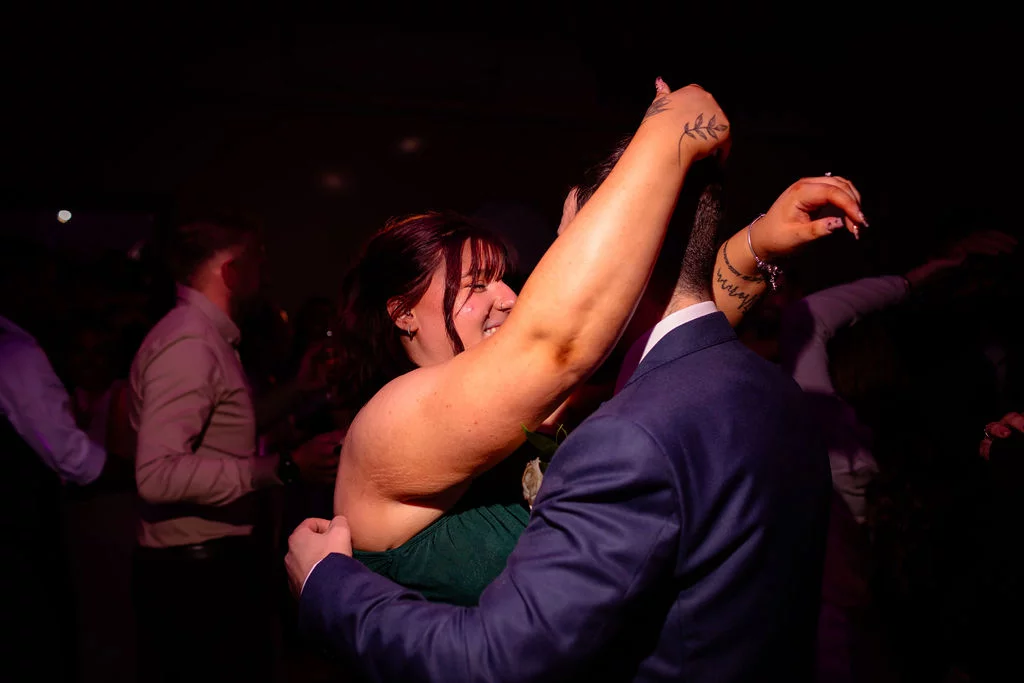 A newlywed couple share a joyful moment dancing together on a dark, ambiently lit evening dance floor surrounded by wedding guests.