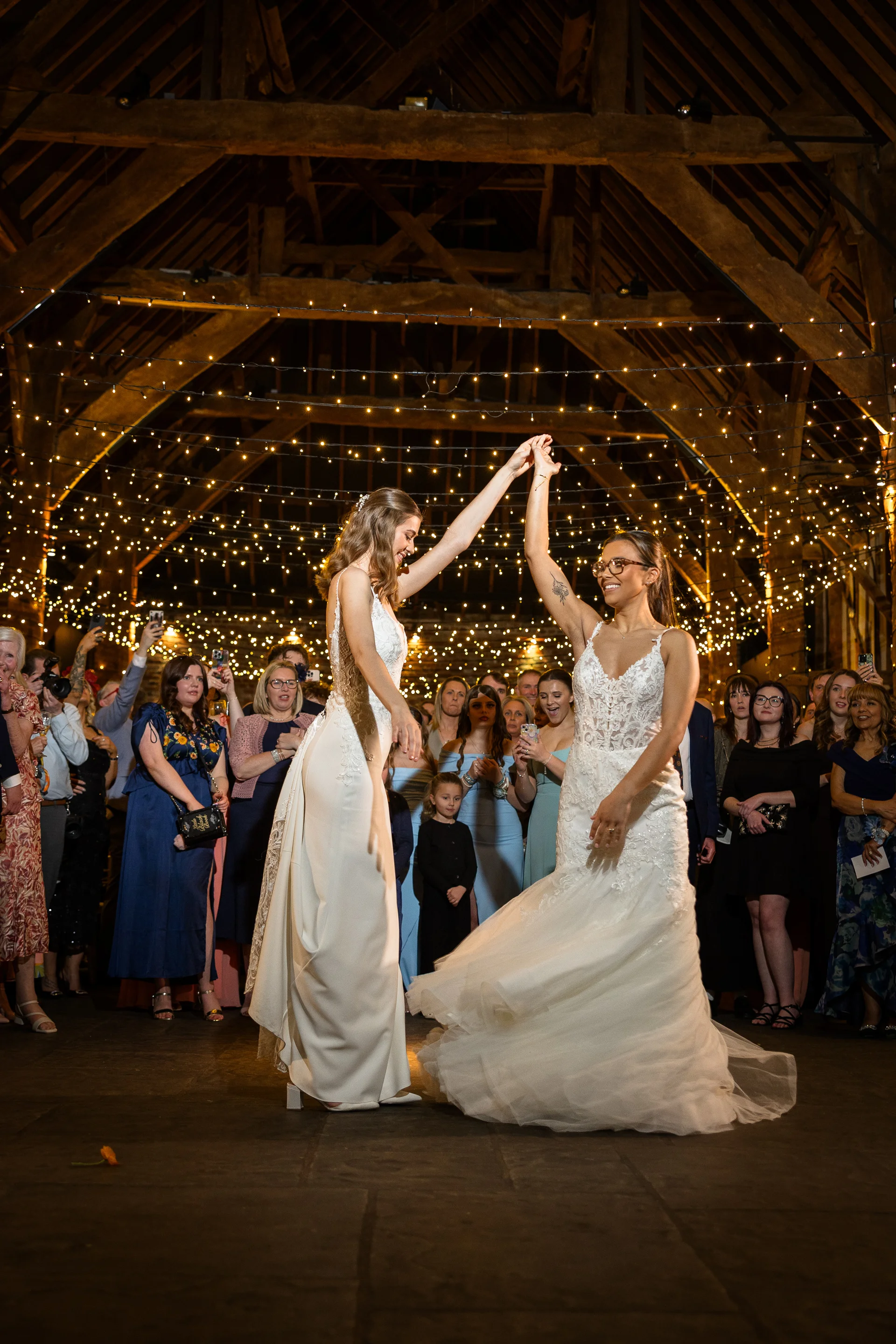 Two brides share their first dance under a canopy of fairy lights inside a rustic oak-framed barn surrounded by smiling guests.