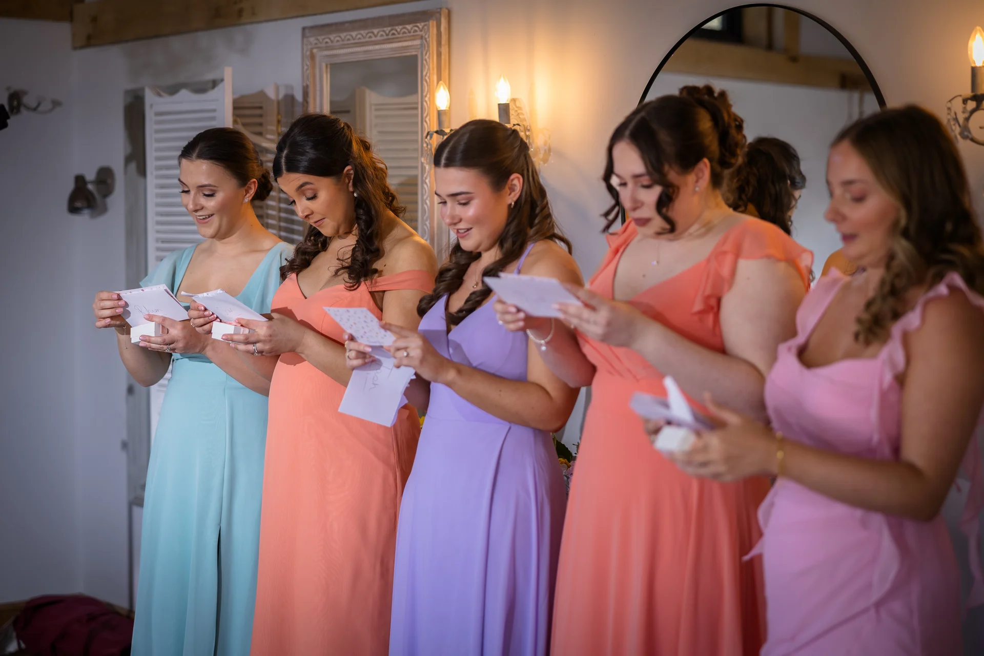 A line of bridesmaids wearing colourful pastel dresses read heartfelt letters together during morning wedding preparations.
