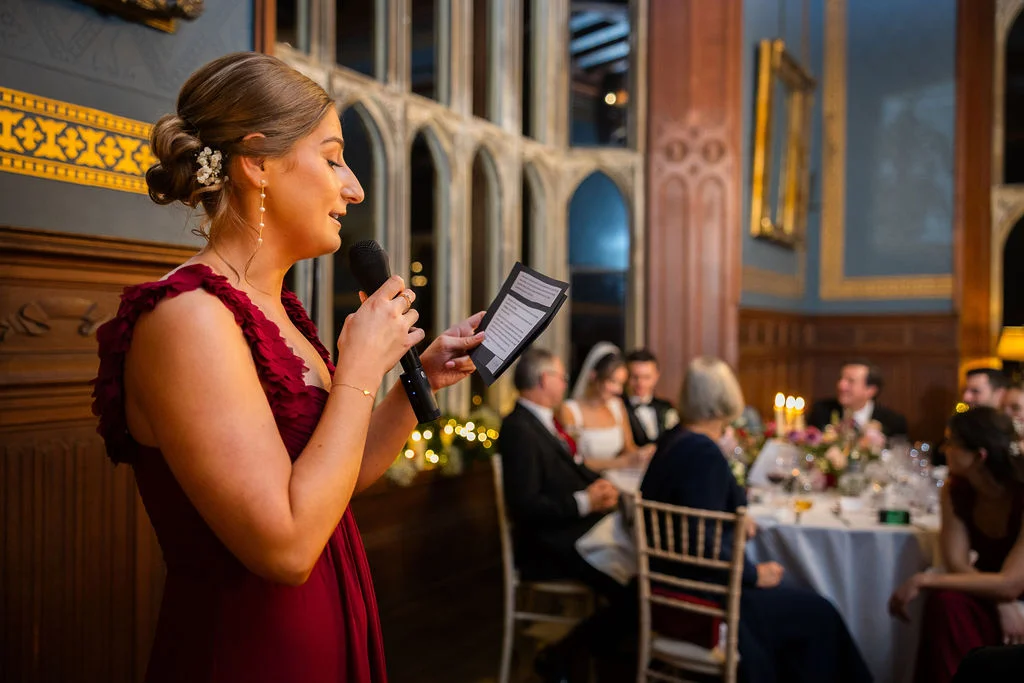 A bridesmaid in a burgundy dress delivers a heartfelt speech holding a microphone and notes in a wood-panelled reception hall.