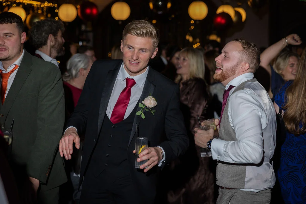 Groomsmen and guests laughing and dancing together holding drinks during a festive evening wedding celebration.