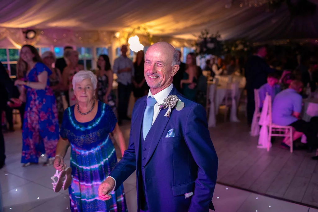 A smiling man in a blue suit and waistcoat dances happily on an illuminated dance floor during a lively evening wedding reception inside a marquee.