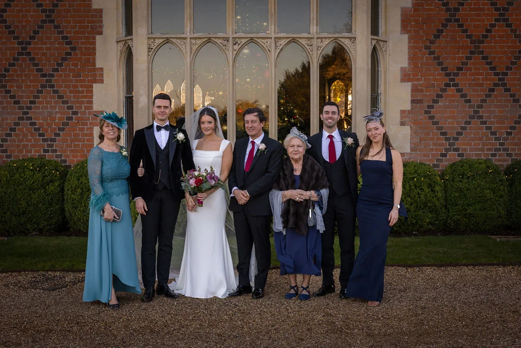 A bridal party and family members pose for a formal group portrait outside a grand red brick manor house with large arched windows.
