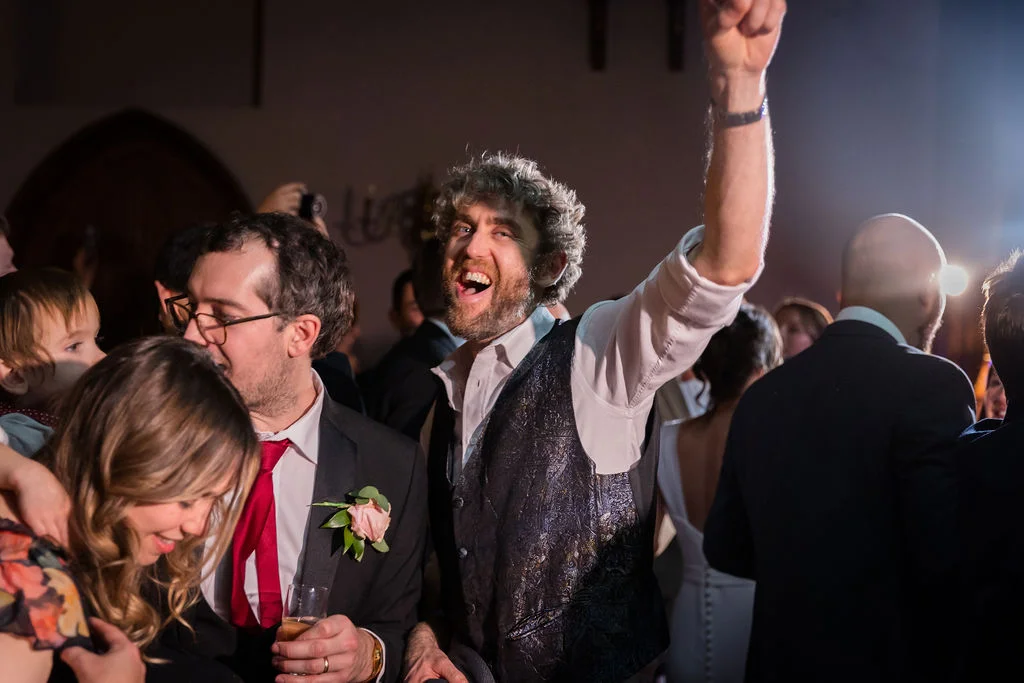 A joyful groom with a patterned waistcoat raises his fist in celebration while cheering and dancing on a lively evening reception dance floor.
