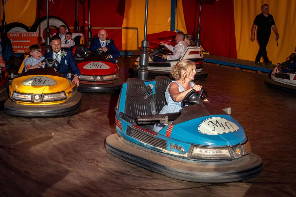 Wedding guests enjoy riding bumper cars inside a colourful retro funfair tent during the evening reception.