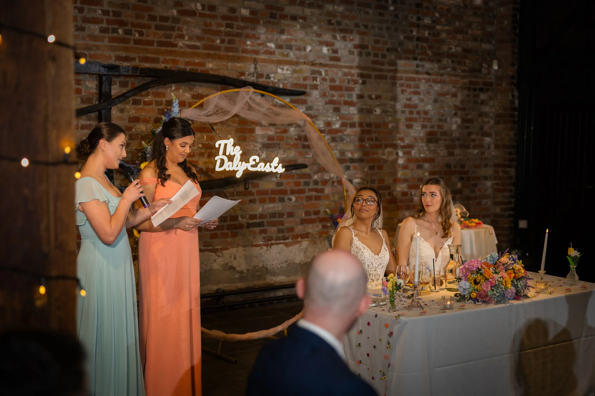 A bridesmaid delivers a speech standing beside the top table where two brides sit attentively in front of an exposed brick wall.