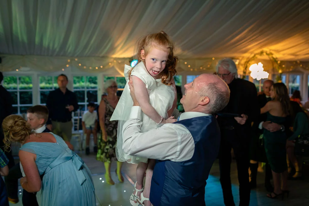 A joyful wedding guest happily lifts a young flower girl into the air on the illuminated dance floor inside a draped marquee.