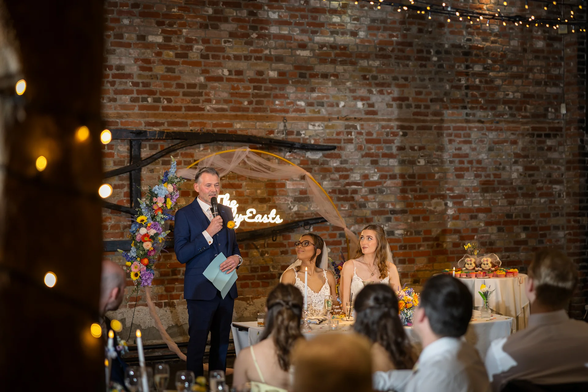 A guest delivers a speech with a microphone at the top table in a rustic brick barn venue as two brides look on and laugh.