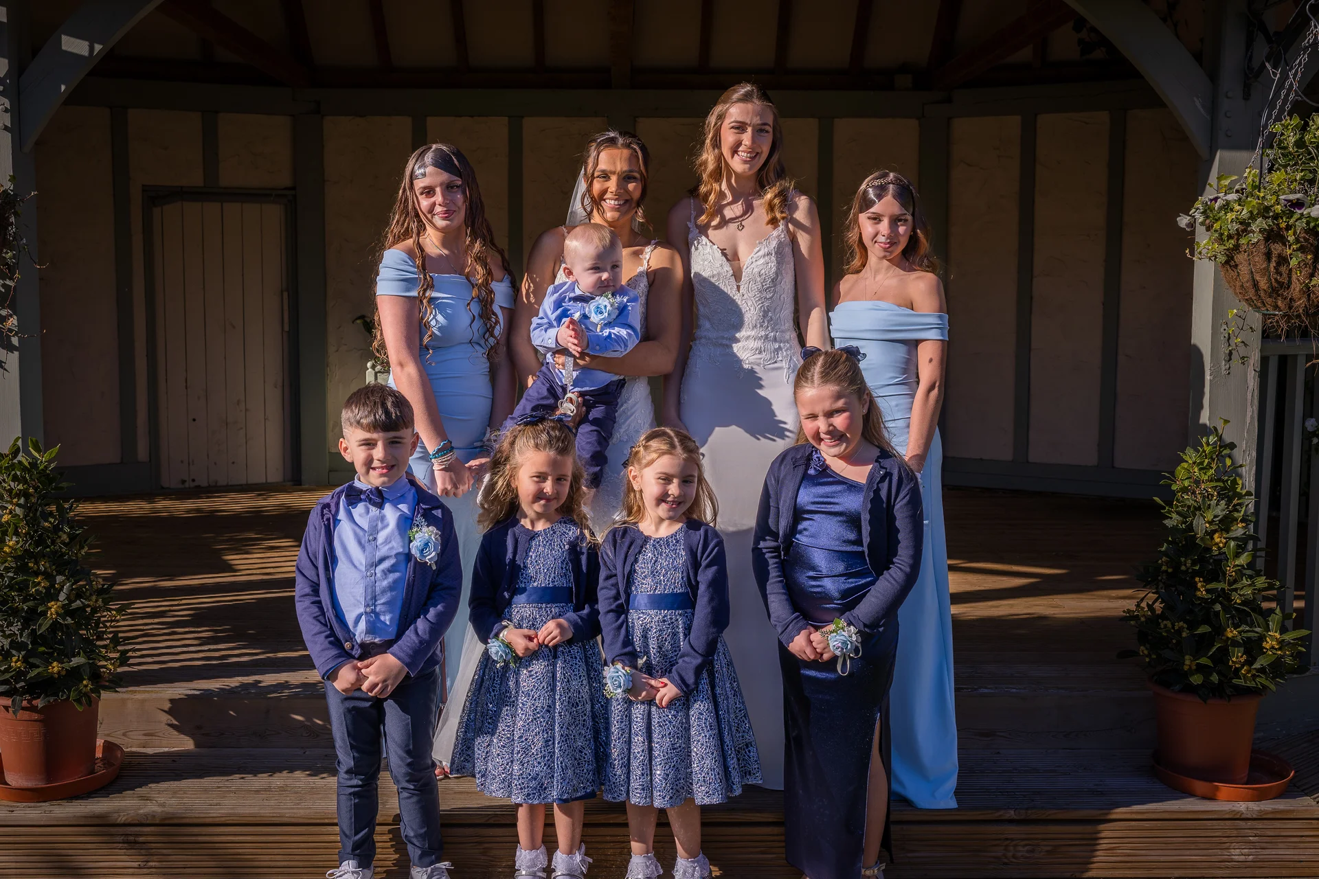 A bride poses outdoors with her bridal party, including matching bridesmaids in blue dresses, young flower girls, page boys, and a baby held in her arms.
