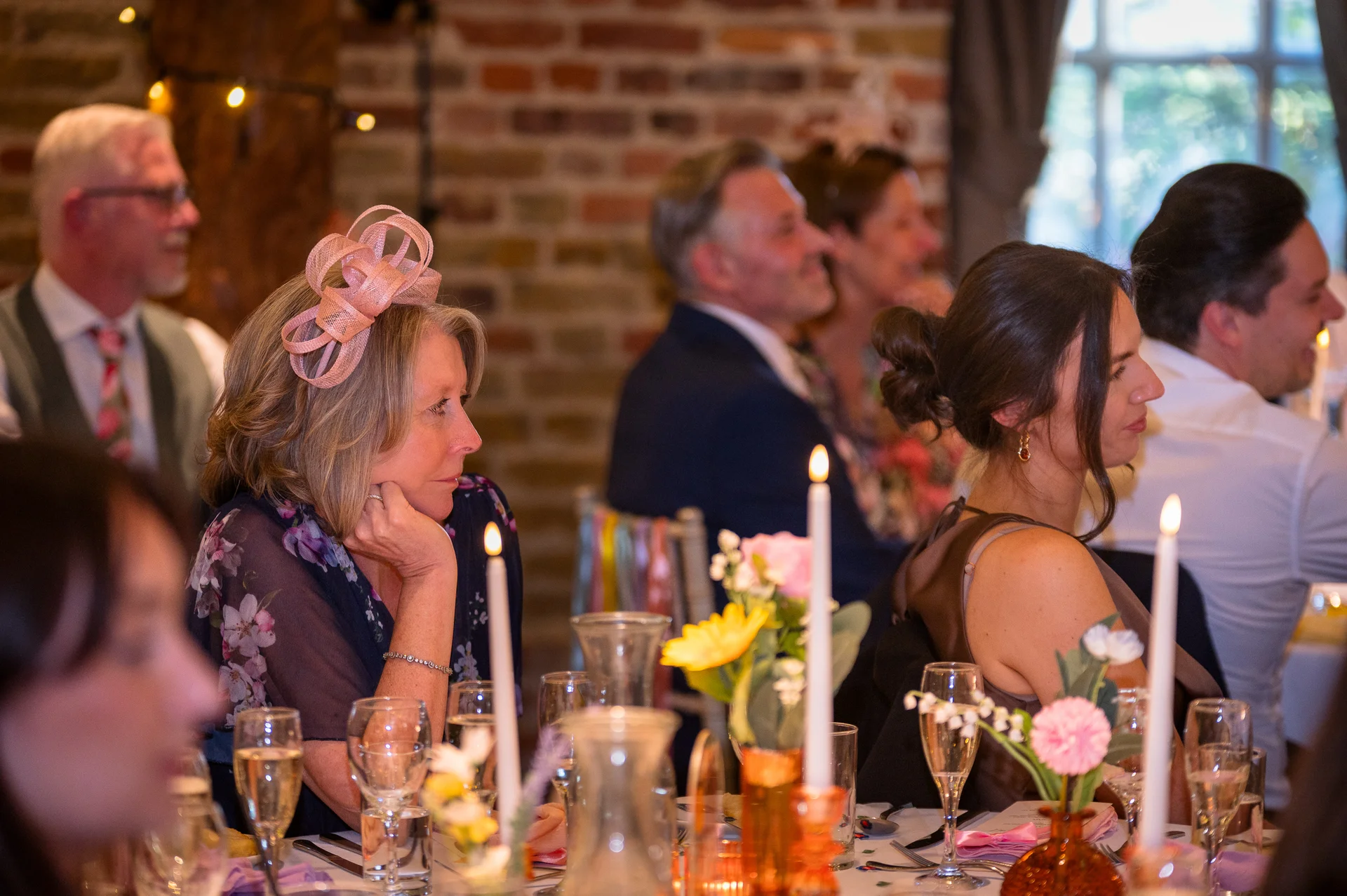 Wedding guests attentively listen to speeches at a rustic barn reception featuring exposed brick walls and a beautifully decorated tablescape with taper candles.