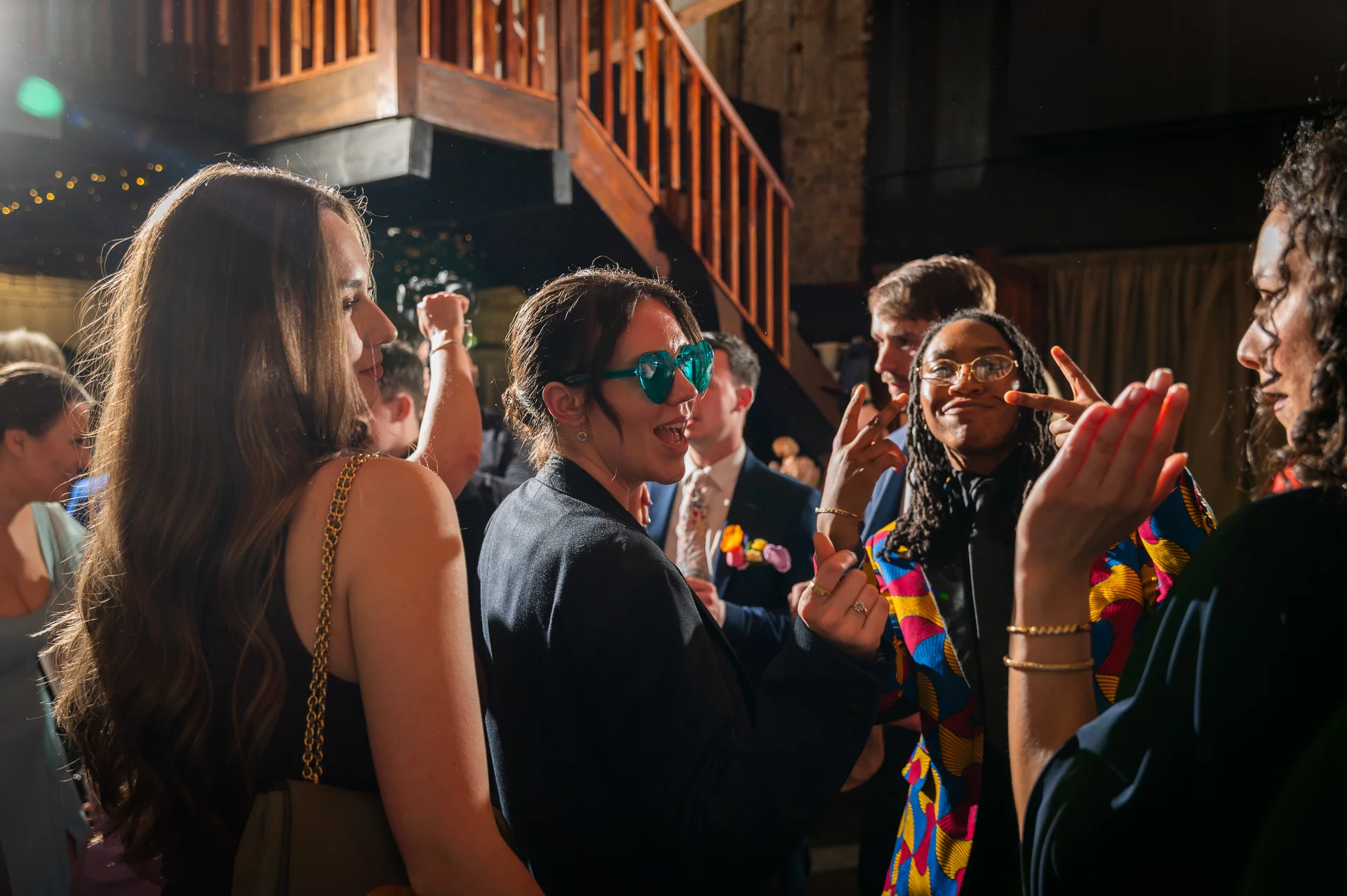 A candid and joyful group of well-dressed wedding guests laugh and celebrate together indoors during an energetic evening reception.