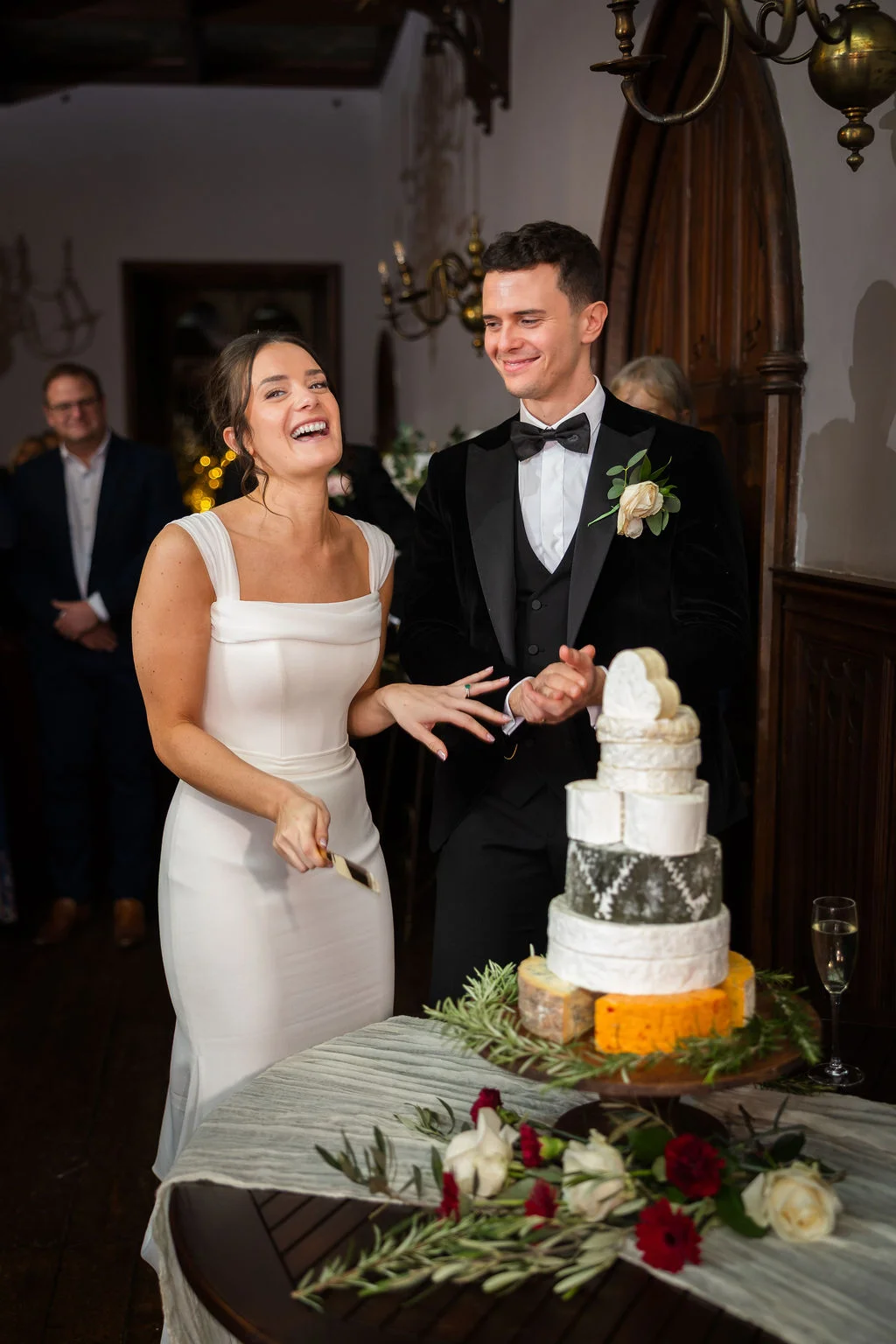 A joyful bride and groom laugh heartily while cutting a tiered cheese wedding tower in a stately dining room.
