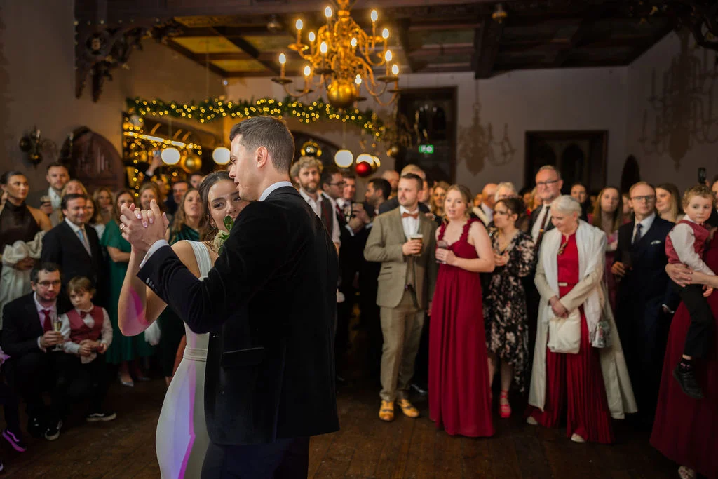 The newlywed couple share their first dance surrounded by a crowd of watching family and friends in a candlelit venue with a grand chandelier.