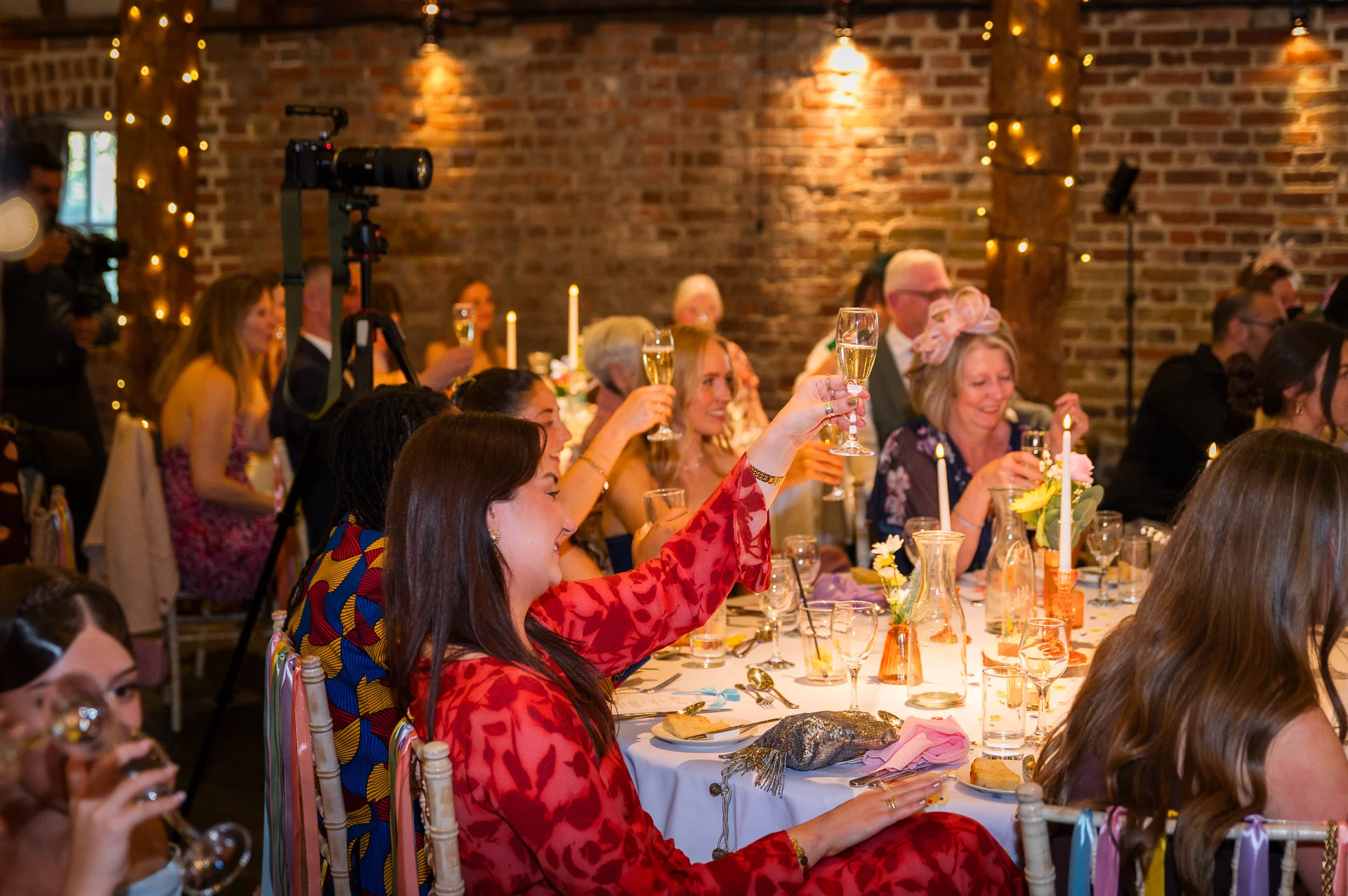 A wedding guest in a red patterned dress raises a glass of champagne in a toast during a lively wedding reception inside a rustic brick barn lit by fairy lights.