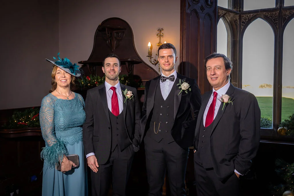 A formal group portrait featuring a mother of the bride in a teal lace dress and matching fascinator alongside three men in tailored black and charcoal suits with red ties and waistcoats inside a grand country house.