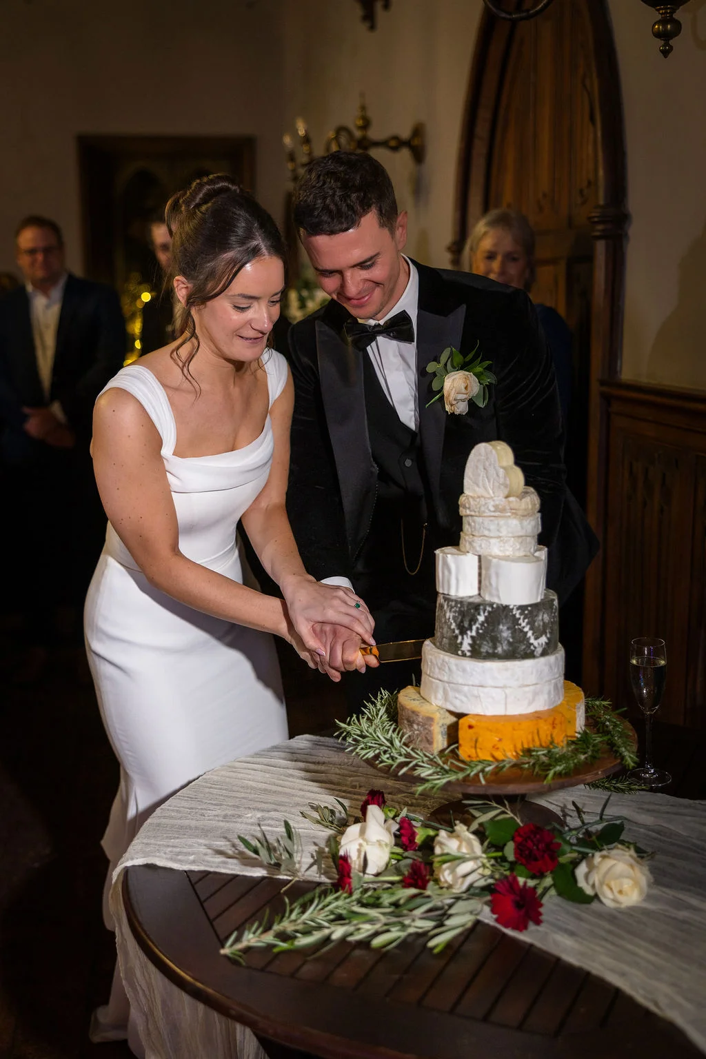 A smiling bride and groom cut a unique tiered cheese wedding tower together at an indoor evening reception.