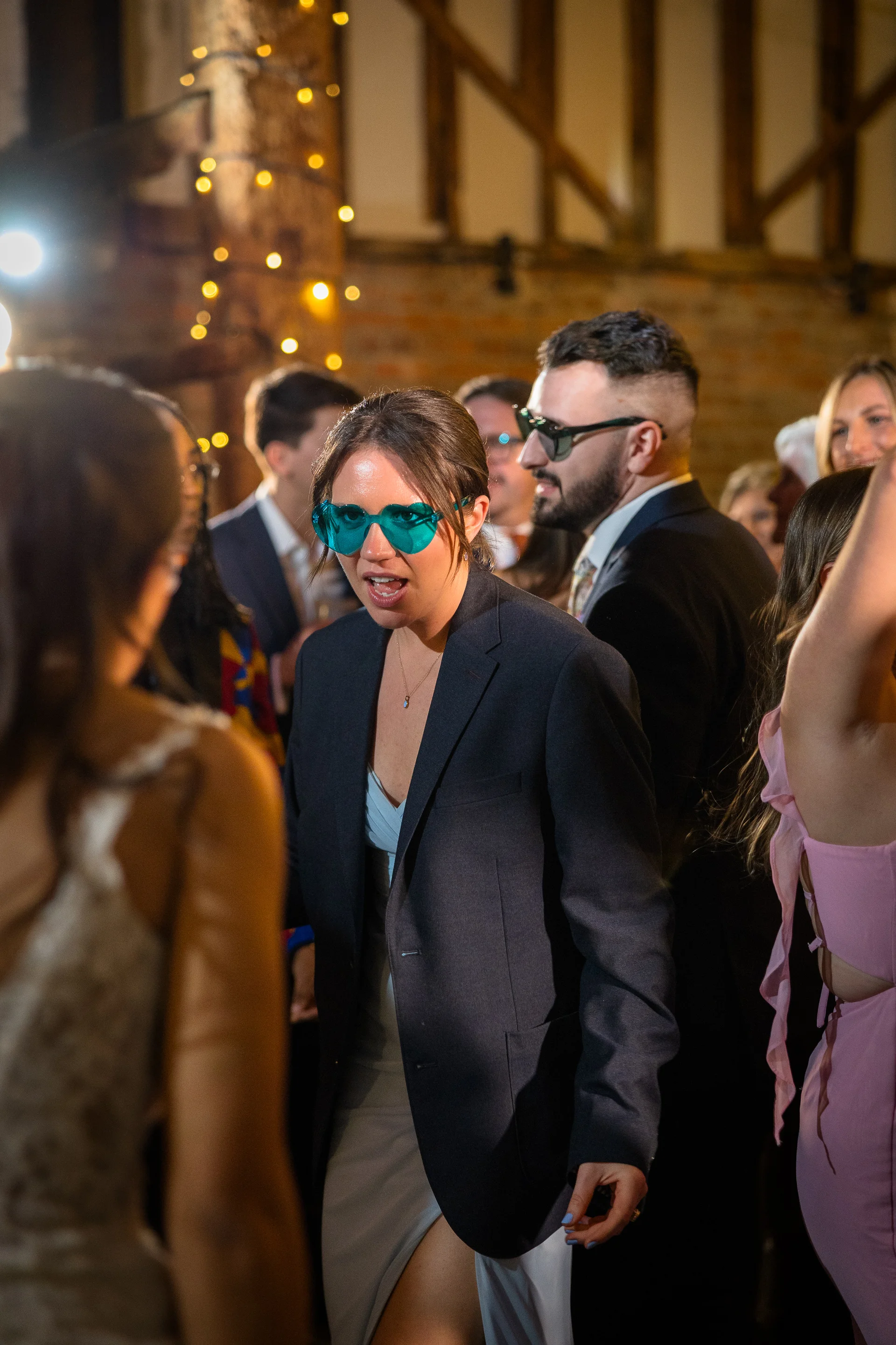 A guest wearing a blazer and blue heart-shaped sunglasses dances joyfully among a lively crowd under the rustic exposed beams of a barn reception.