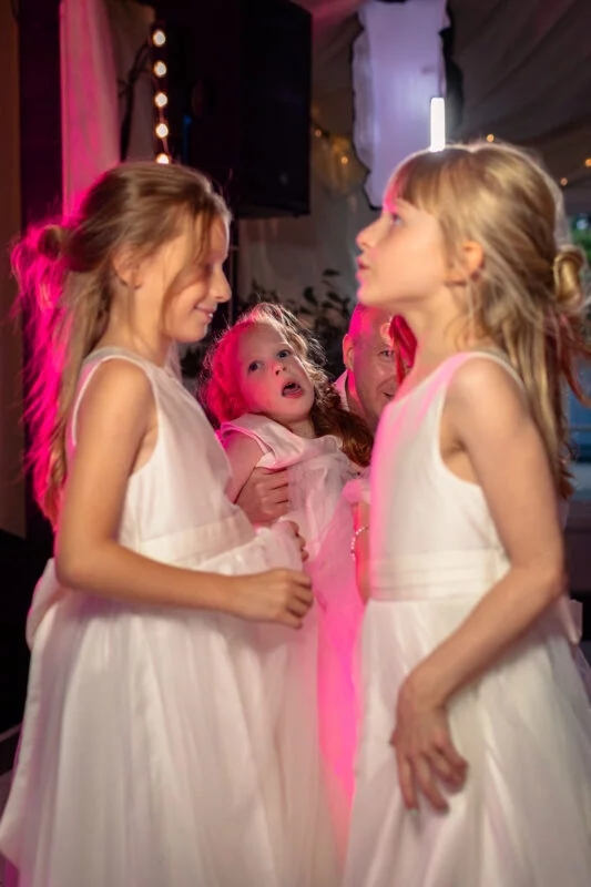 Several young flower girls in matching white dresses enjoy the wedding reception under colourful stage lighting.