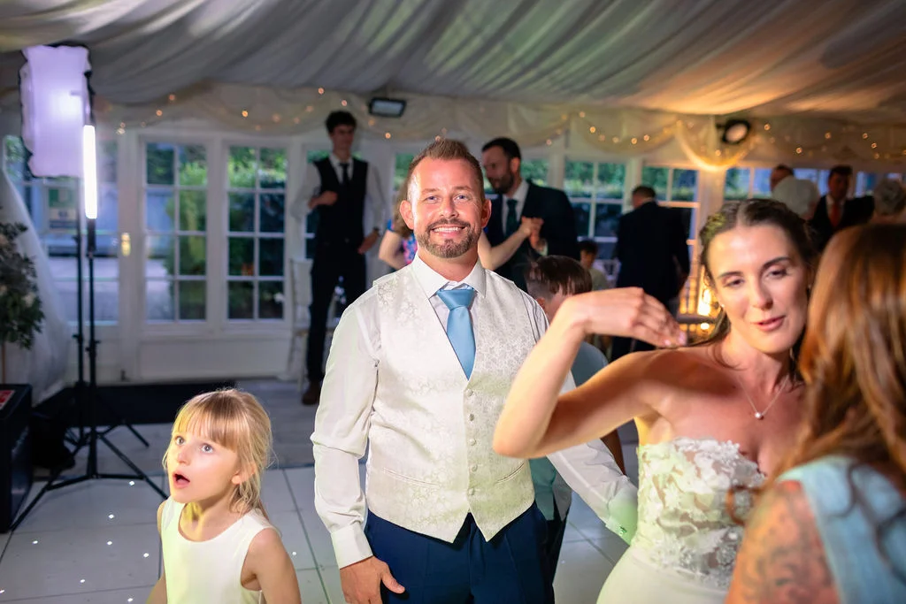 A smiling groom in a waistcoat and blue tie stands on a lit dance floor during a marquee wedding reception, accompanied by a young flower girl and guests celebrating.