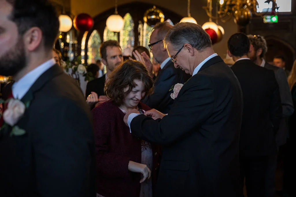 A wedding guest pins a buttonhole onto another guest during a lively indoor reception gathering.