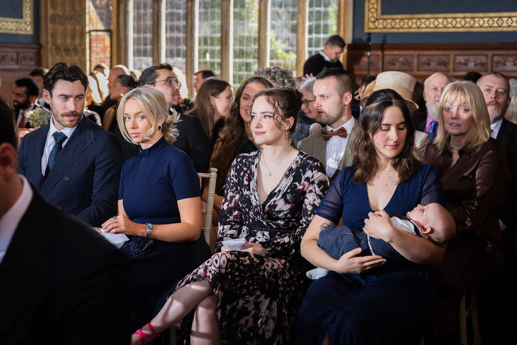A group of well-dressed wedding guests are seated attentively inside a historic venue with large windows, including a woman holding a baby.