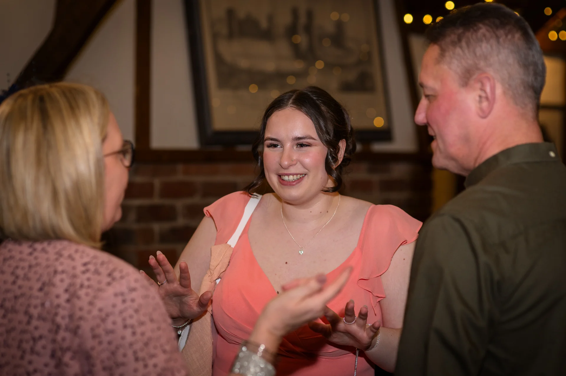 A smiling guest wearing a coral dress chats animatedly with friends inside a rustic venue featuring exposed brickwork.