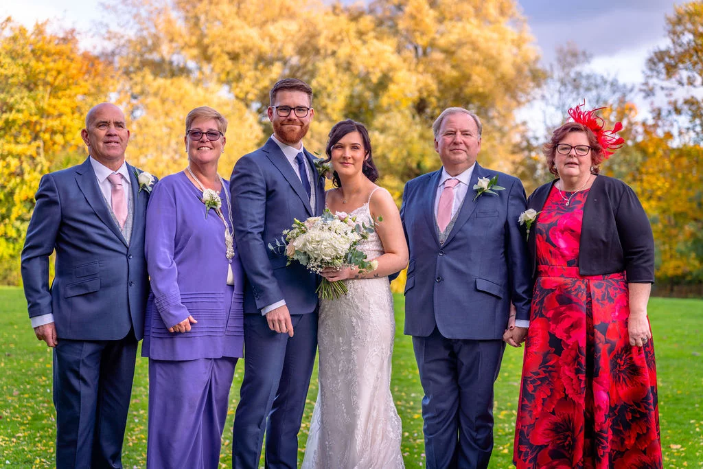 A newlywed couple and family members pose together outdoors on a lush green lawn against a vibrant backdrop of golden autumn trees.